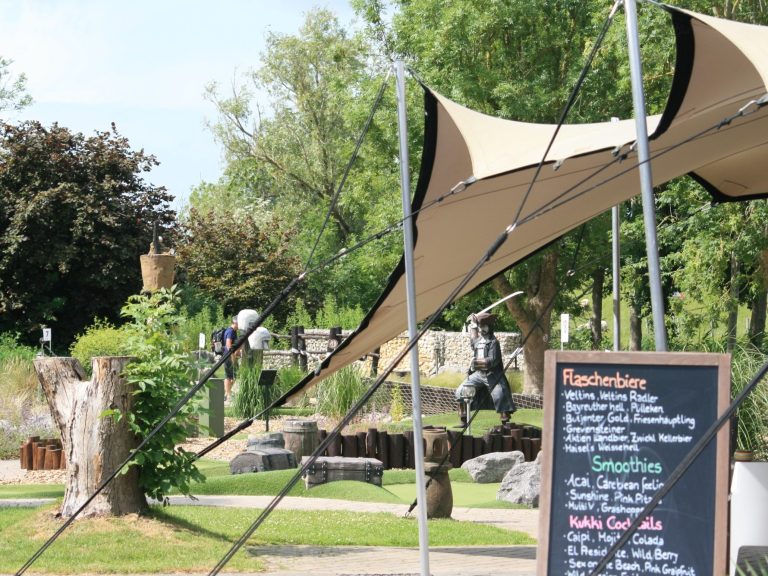 Minnigolf Spielplatz mit Sonnenschutz, Tafel mit Menu, Bäume im Hintergrund und mittelalterliche Statue.